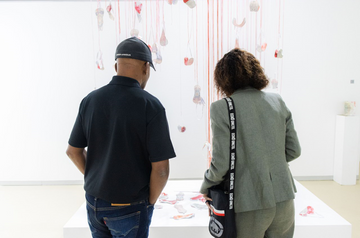 Gallerygoers viewing a suspended mixed-media installation hanging over a low white platform.