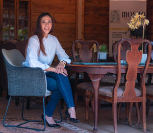 Portrait photograph of a smiling woman with long brown hair, wearing a white button-up blouse, blue jeans, and black pointed heels. She sits cross-legged in a grey armchair beside a polished wooden dining table inside a warm, wooden-paneled room.