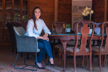 Portrait photograph of a smiling woman with long brown hair, wearing a white button-up blouse, blue jeans, and black pointed heels. She sits cross-legged in a grey armchair beside a polished wooden dining table inside a warm, wooden-paneled room.